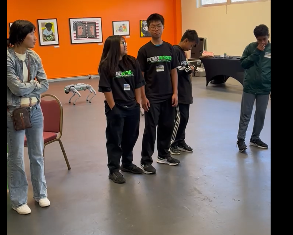 Student builders in RoboRoots shirts standing in a line as a quadruped robot walks behind them at AAACC