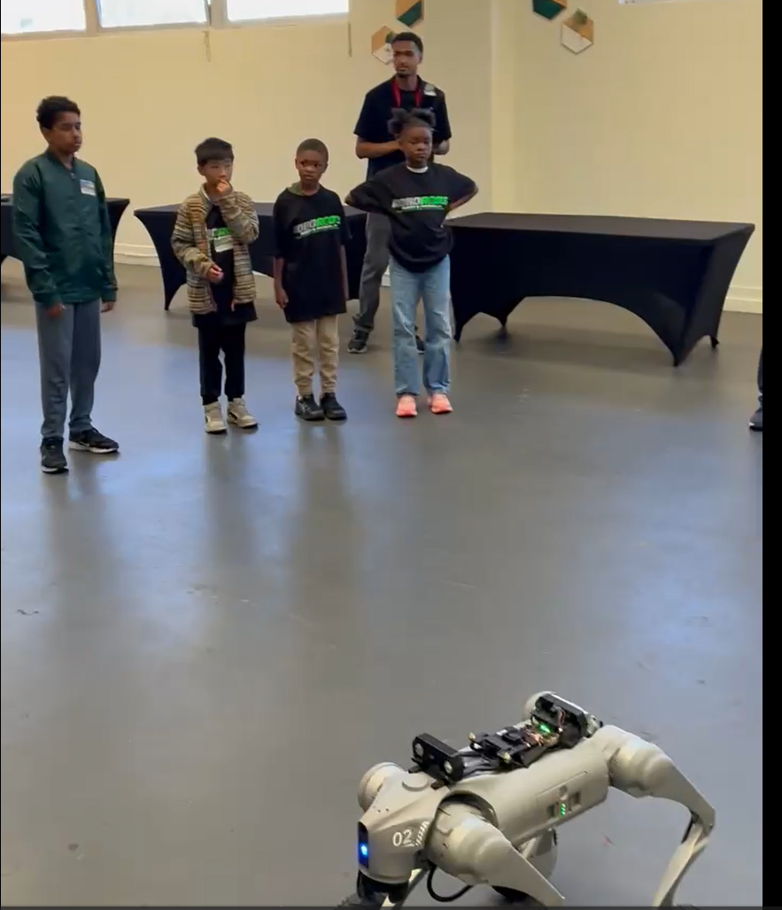 Young builders and a mentor watching a quadruped robot move toward them across the arena floor